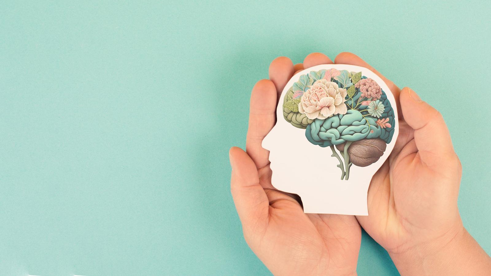 Hands set against a pastel teal background hold an artistic cutout of a human head with a brain